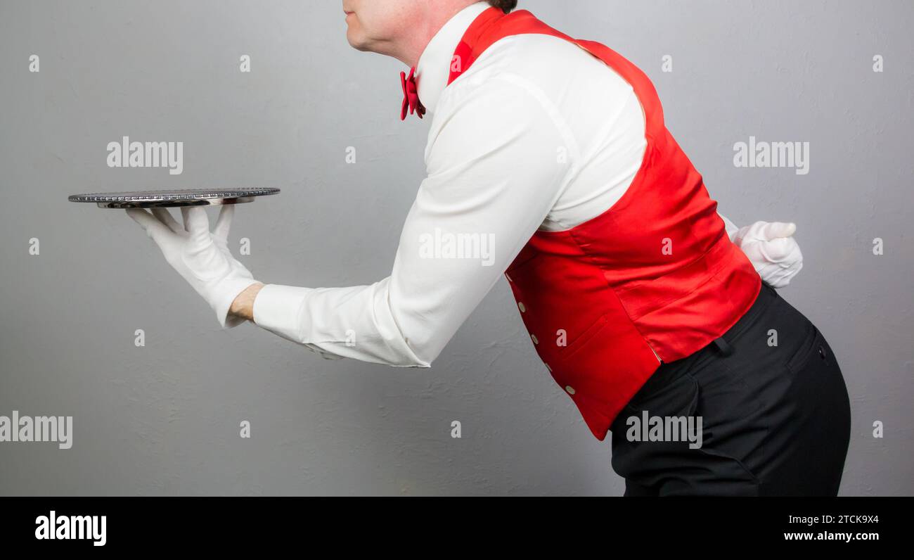 Portrait of Waiter in Red Vest or Waistcoat and Red Bow Tie Holding Silver Serving Tray. Concept of Service Industry and Professional Hospitality. Stock Photo