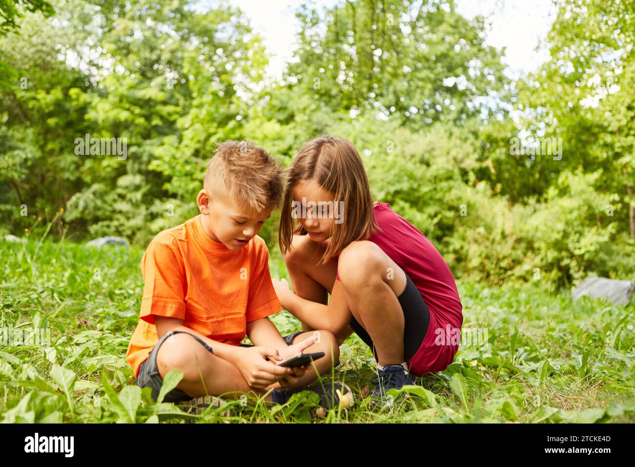 Girl crouching next to boy using smart phone while sitting on grass at park Stock Photo