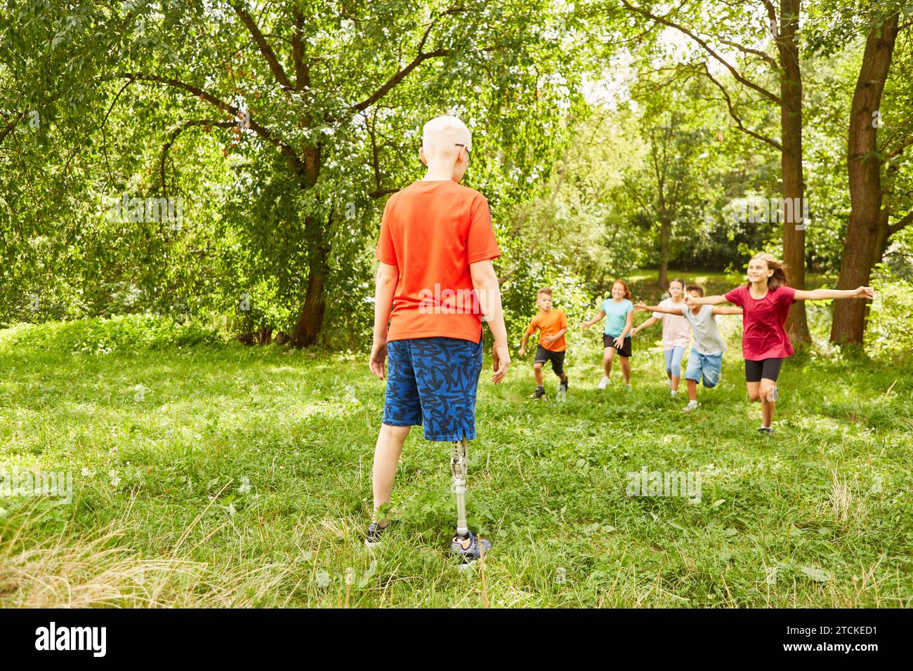 Happy kids running towards disabled male friend standing on grass at park Stock Photo