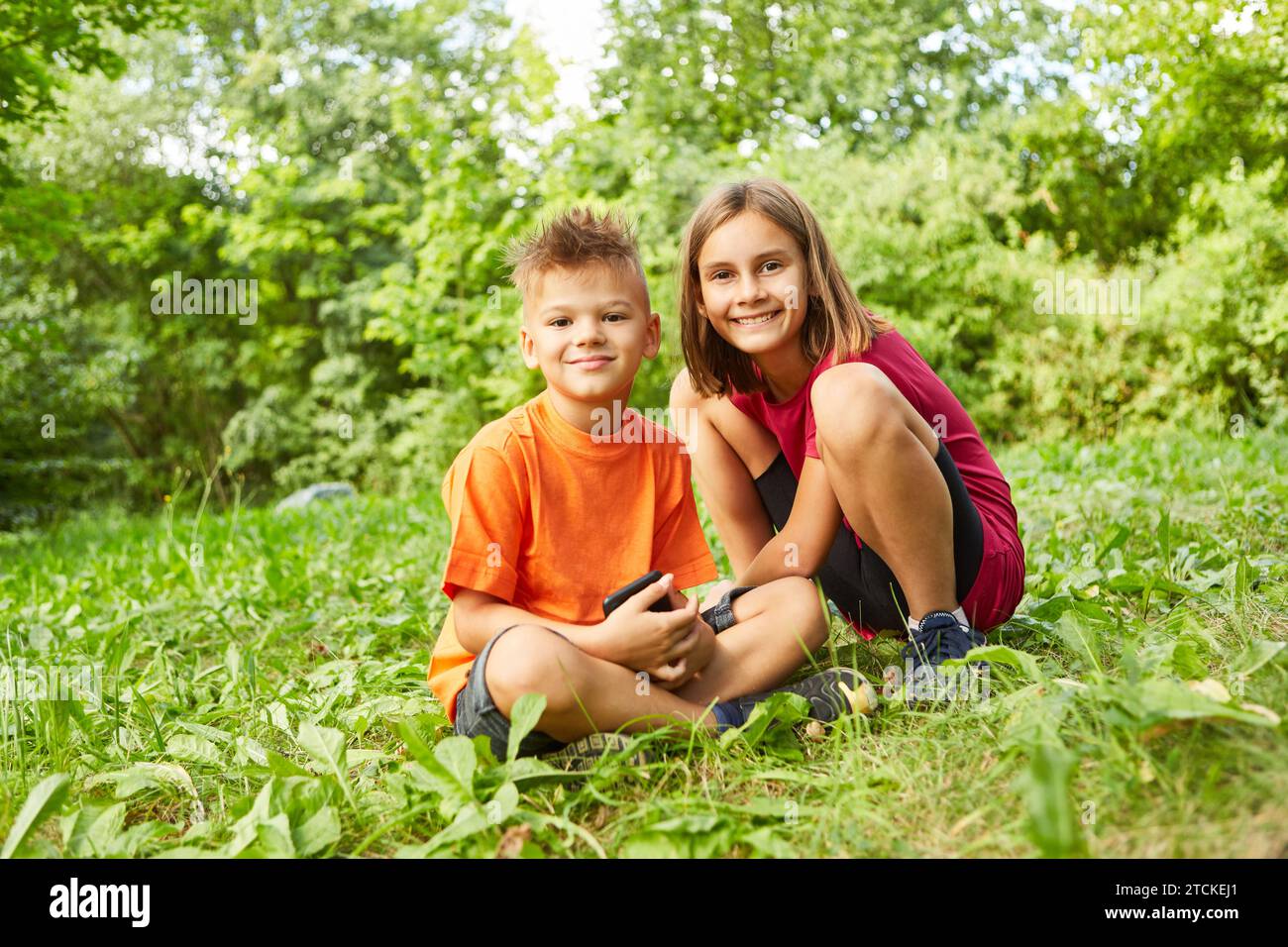 Portrait of smiling girl crouching next to boy sitting with smart phone at park Stock Photo