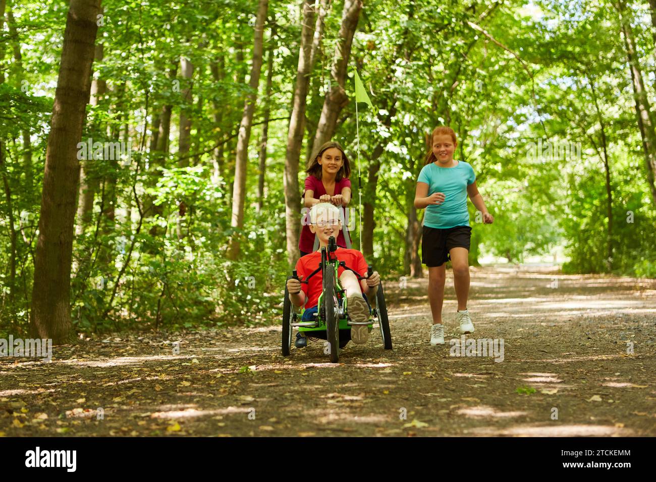 Playful girl running with disabled boy riding recumbent bike on footpath at forest Stock Photo