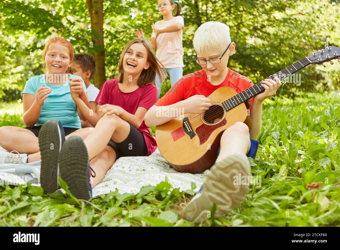 Happy female friends sitting next to boy playing guitar while sitting on blanket at park Stock Photo