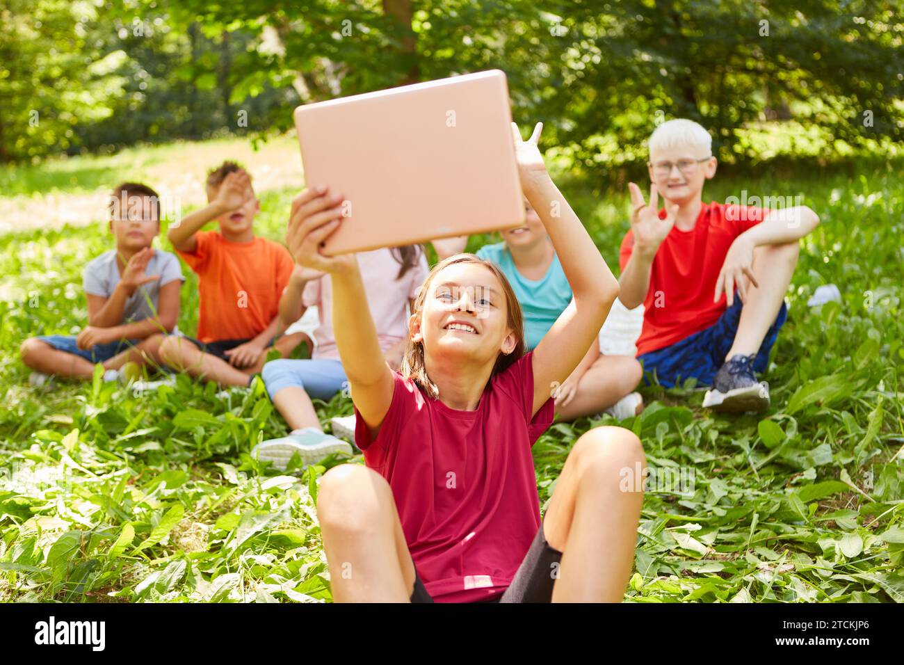 Happy girl gesturing and taking selfie with friends through tablet PC while sitting at park Stock Photo