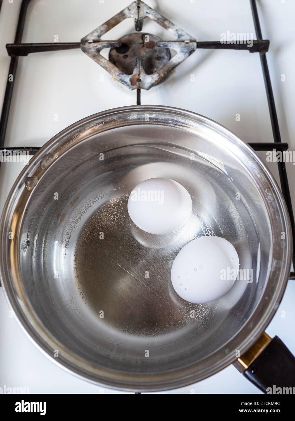 two white chicken eggs are boiling in steel ladle on gas stove close up Stock Photo