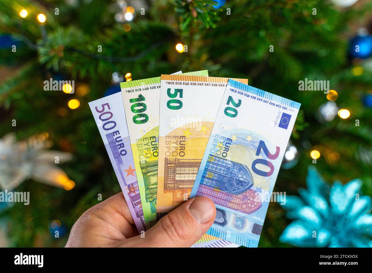 Augsburg, Bavaria, Germany - December 13, 2023: Hand holding cash euro banknotes in front of a decorated Christmas tree. Costs and gift concept *** Hand hält Bargeld Euro Geldscheine vor einen geschmückten Weihnachtsbaum. Kosten und Geschenk Konzept Stock Photo