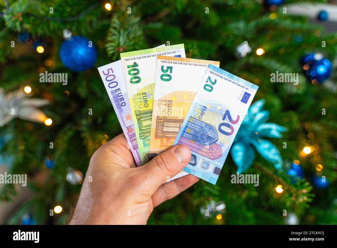 Augsburg, Bavaria, Germany - December 13, 2023: Hand holding cash euro banknotes in front of a decorated Christmas tree. Costs and gift concept *** Hand hält Bargeld Euro Geldscheine vor einen geschmückten Weihnachtsbaum. Kosten und Geschenk Konzept Stock Photo