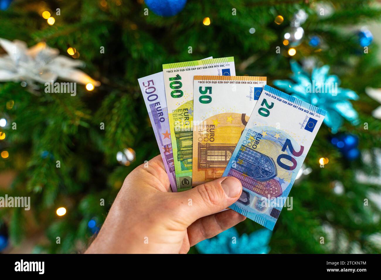 Augsburg, Bavaria, Germany - December 13, 2023: Hand holding cash euro banknotes in front of a decorated Christmas tree. Costs and gift concept *** Hand hält Bargeld Euro Geldscheine vor einen geschmückten Weihnachtsbaum. Kosten und Geschenk Konzept Stock Photo