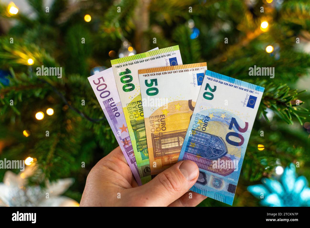 Augsburg, Bavaria, Germany - December 13, 2023: Hand holding cash euro banknotes in front of a decorated Christmas tree. Costs and gift concept *** Hand hält Bargeld Euro Geldscheine vor einen geschmückten Weihnachtsbaum. Kosten und Geschenk Konzept Stock Photo