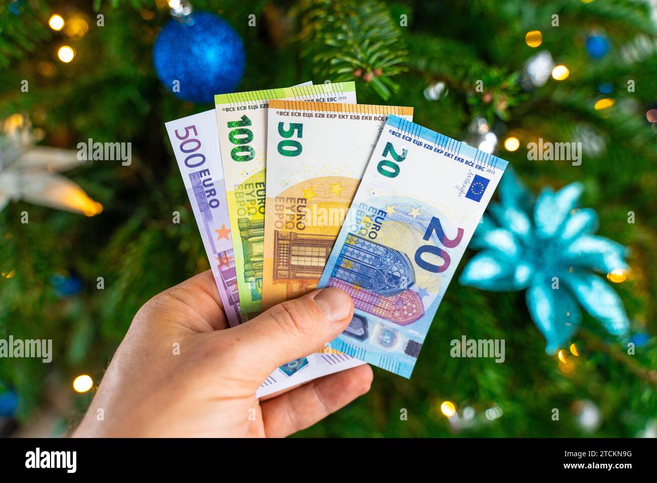 Augsburg, Bavaria, Germany - December 13, 2023: Hand holding cash euro banknotes in front of a decorated Christmas tree. Costs and gift concept *** Hand hält Bargeld Euro Geldscheine vor einen geschmückten Weihnachtsbaum. Kosten und Geschenk Konzept Stock Photo