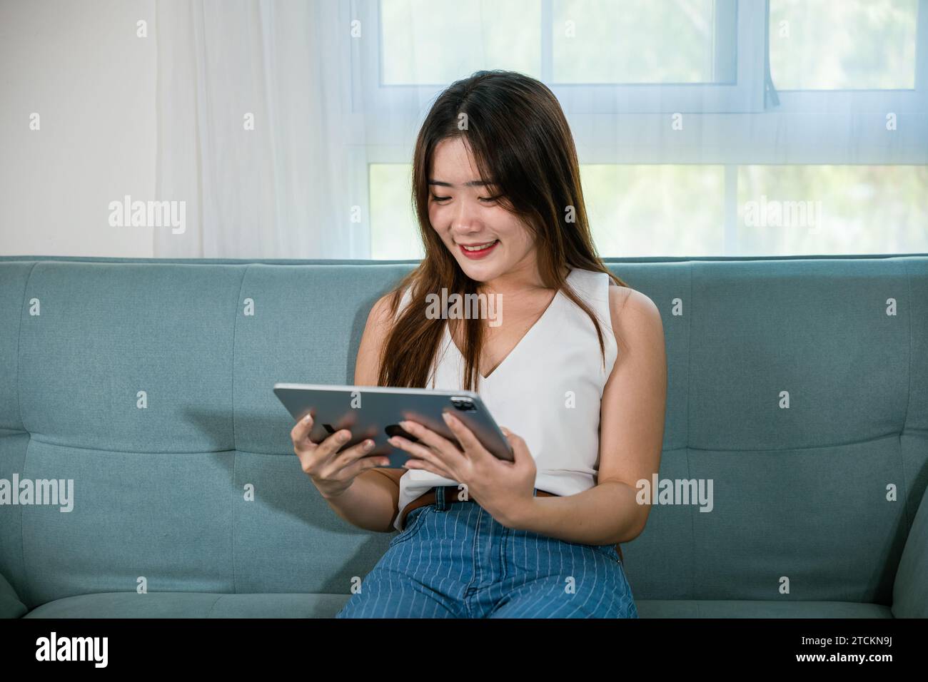 Happy Asian woman playing smart tablet at home while relaxing on sofa ...