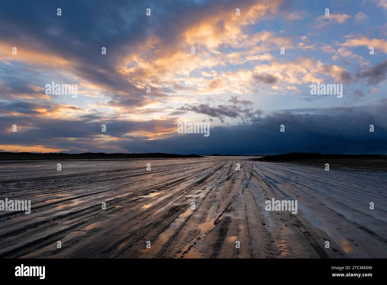 Traces of car tires on a muddy beach, leading to the horizon at nightfall, under a beautifully colored sky, threatening clouds in the distance Stock Photo