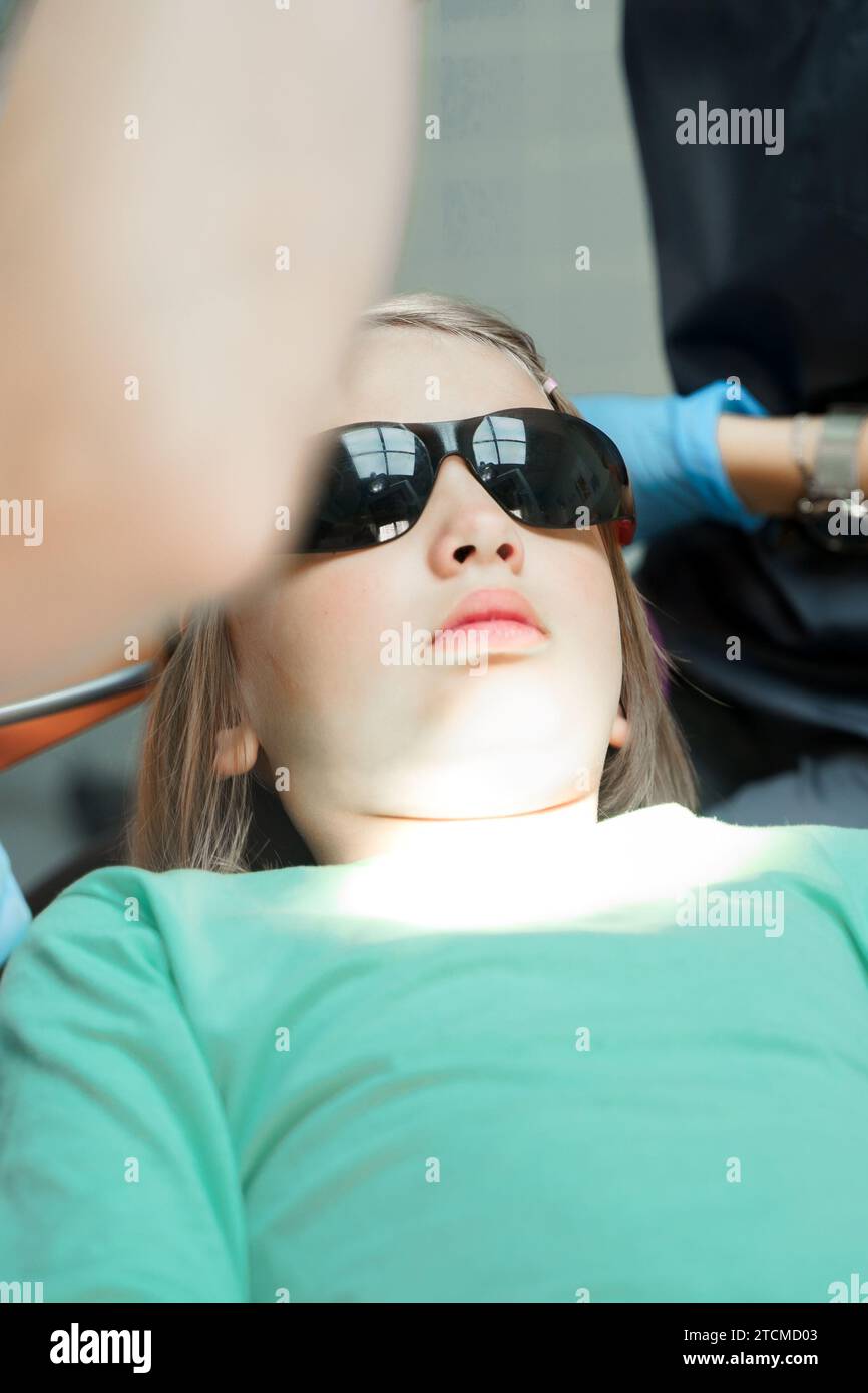 Little scared girl sitting in chair in dentist doctor office. Protective safety sunglasses. Kid,child afraid of tooth extraction, teeth treatment. Ope Stock Photo