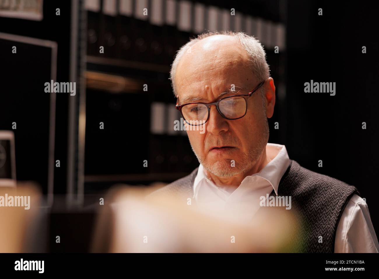 Tired investigator reading confidential federal files, working overhours at criminal case in evidence room. Elderly private detective analzing crime scene report, planning strategy to catch suspect Stock Photo