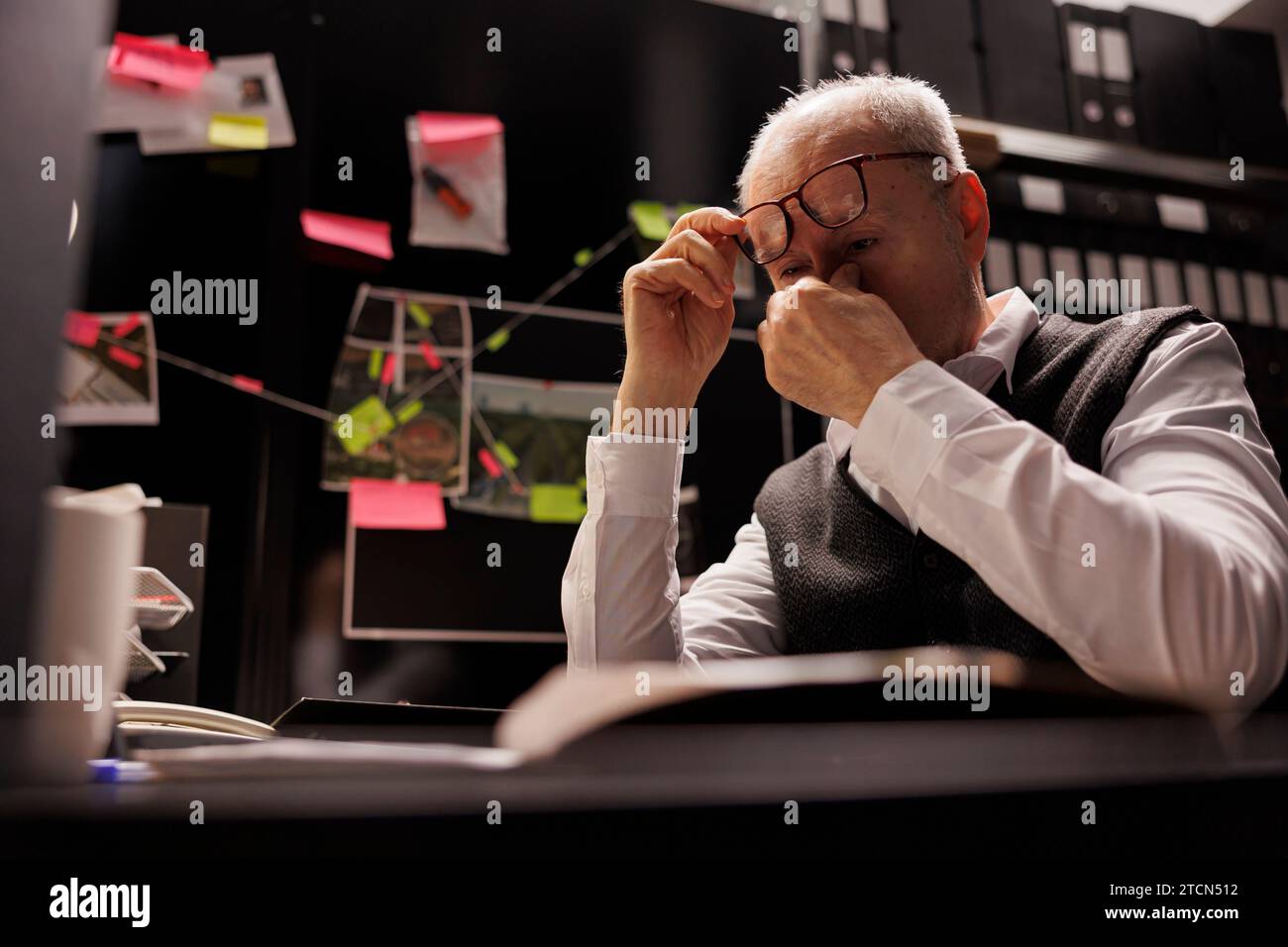 Overworked old investigator analyzing criminology report, working late at night at criminal case in arhive room. Tired private detective checking victim files, looking at crime scene evidence Stock Photo