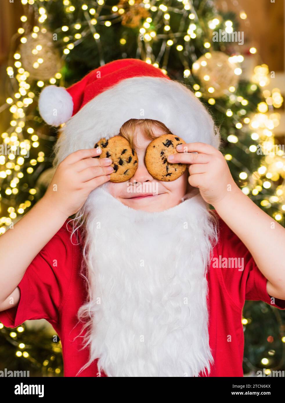 Happy Santa Claus - cute boy child eating a cookie and drinking glass of milk at home Christmas interior. Little Santa picking cookie and glass of Stock Photo