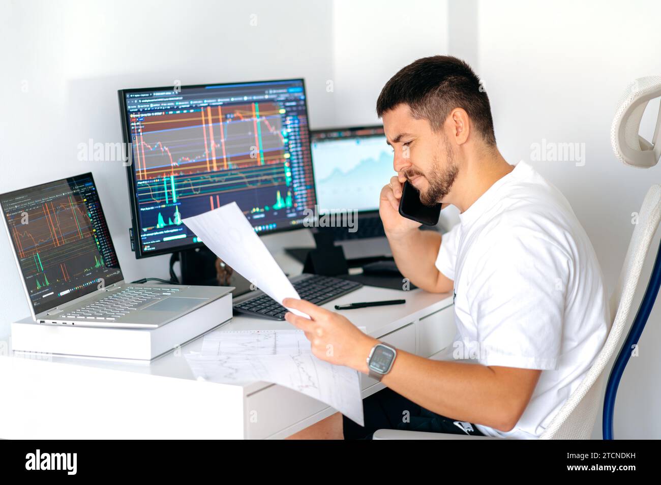 Busy caucasian male crypto trader sitting at home at his work desk in front  of monitors with graphs, talking on the smart phone with colleague,  discussing the prospects of cryptocurrency, profit Stock