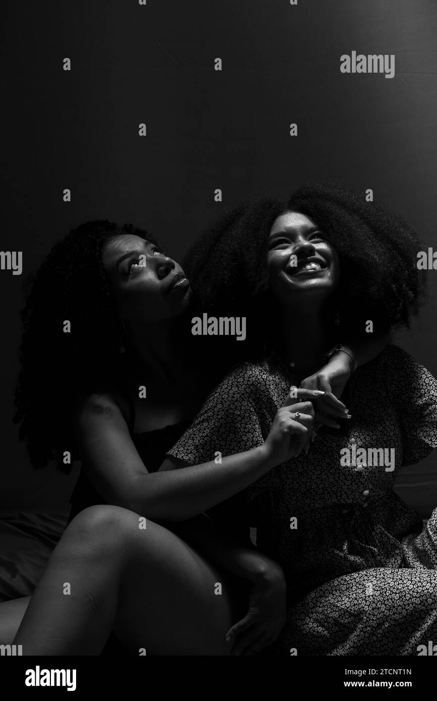 Black and white portrait of two young and beautiful friends sitting, looking up smiling. Studio low light. Solid friendship concept. Stock Photo