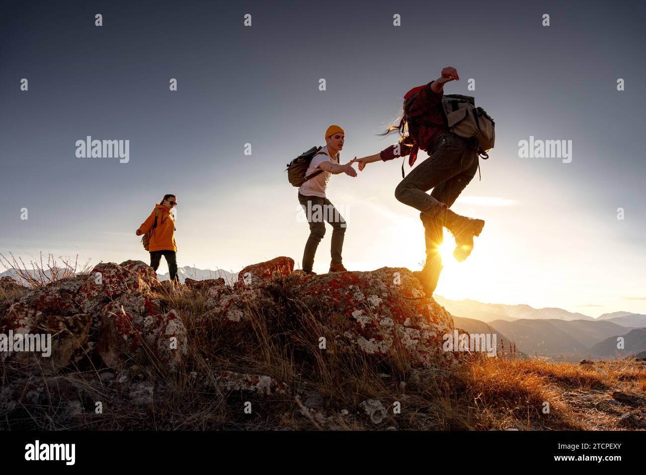 Group of young hikers with backpacks walks with backpacks and helps each other in climbing in sunset mountains Stock Photo