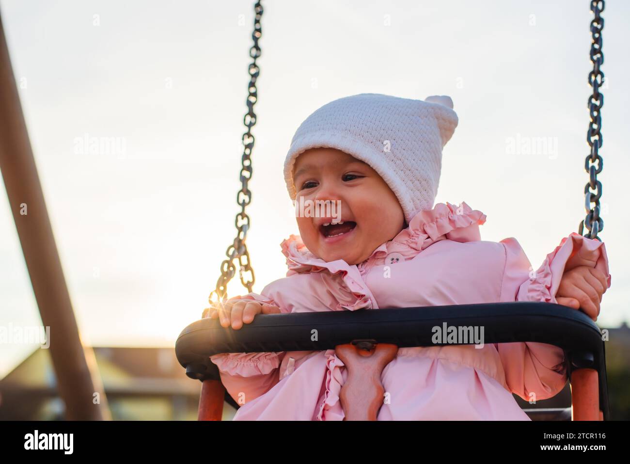 Adorable baby girl with big beautiful eyes and a beanie having fun on a swing ride at a playground in a sunny park Stock Photo