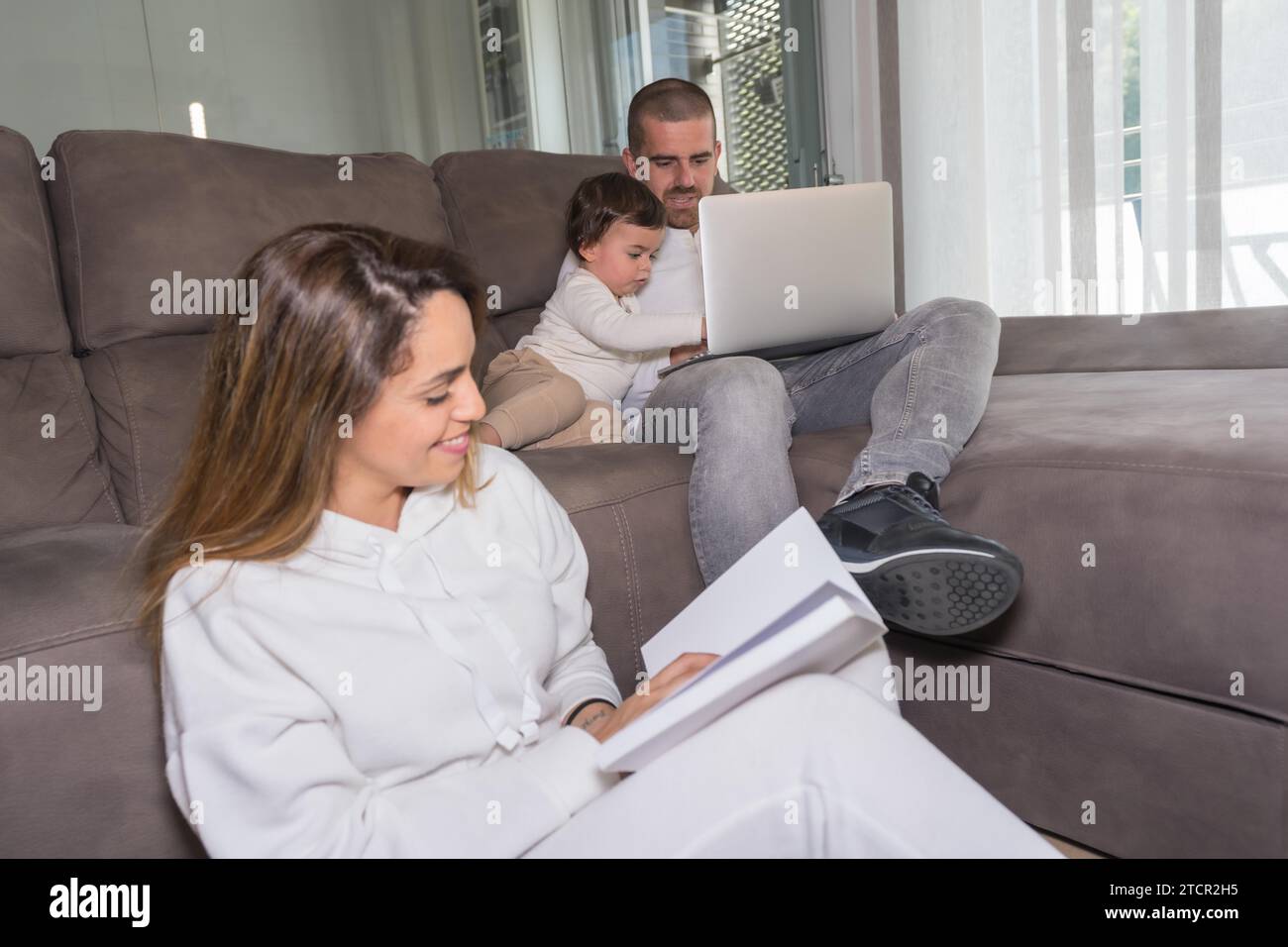 Mother is reading a book while a father is using laptop sitting on the sofa during weekend activities of a family with one child at home Stock Photo