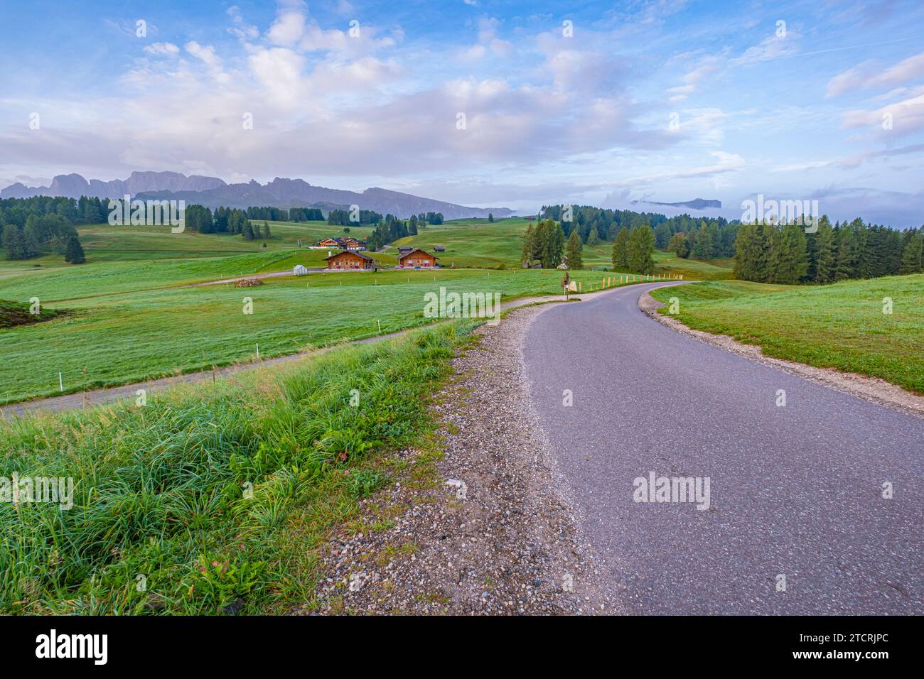 Morning scene, Wood houses dot a green meadow, framed by Alpe di Siusi in the background, under a partly cloudy sky and the calming hues of blue Stock Photo