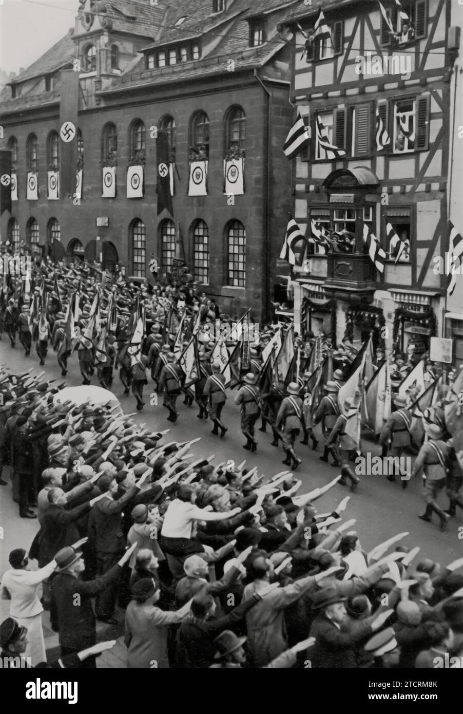 At the 1935 Nuremberg Rally, on the Day of the Armed Forces, soldiers march down the street, carrying the flags of Germany's historic army, as onlookers and supporters salute with their right arm. This powerful scene underscores the military heritage and tradition, showcasing the continuity and historical prestige of the German Army, while also reflecting the widespread public support and participation in Nazi propaganda. Stock Photo