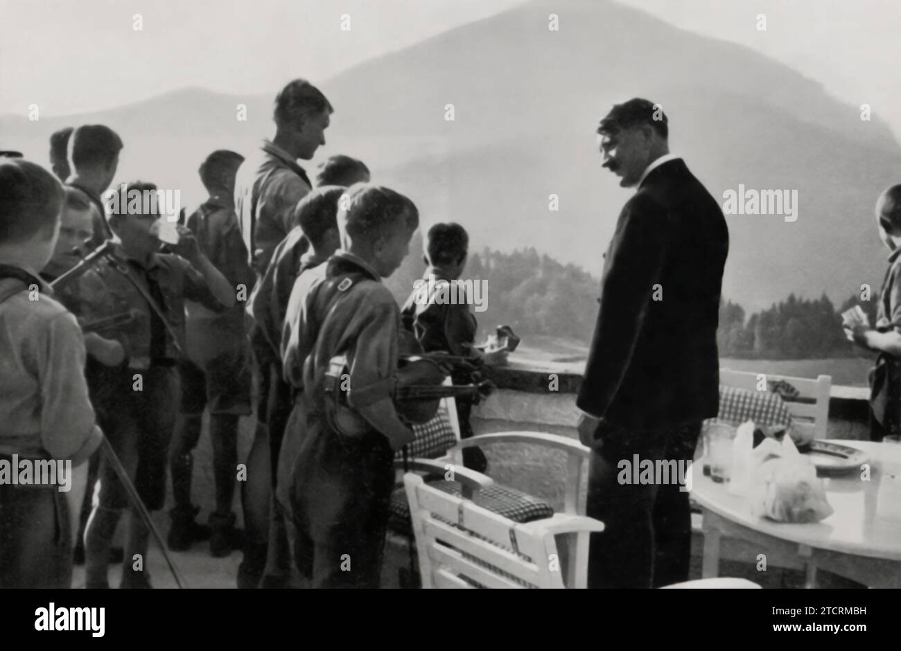 Adolf Hitler is seen with members of the Hitler Youth outside his residence at the Obersalzberg, set against the scenic backdrop of the mountains. This image not only highlights the close involvement of the youth with the Nazi leadership but also the regime's use of picturesque settings to enhance its propaganda. The Obersalzberg, a retreat for high-ranking officials, becomes a stage for showcasing the indoctrination of the younger generation, symbolizing their integration into the core of Nazi ideology. Stock Photo