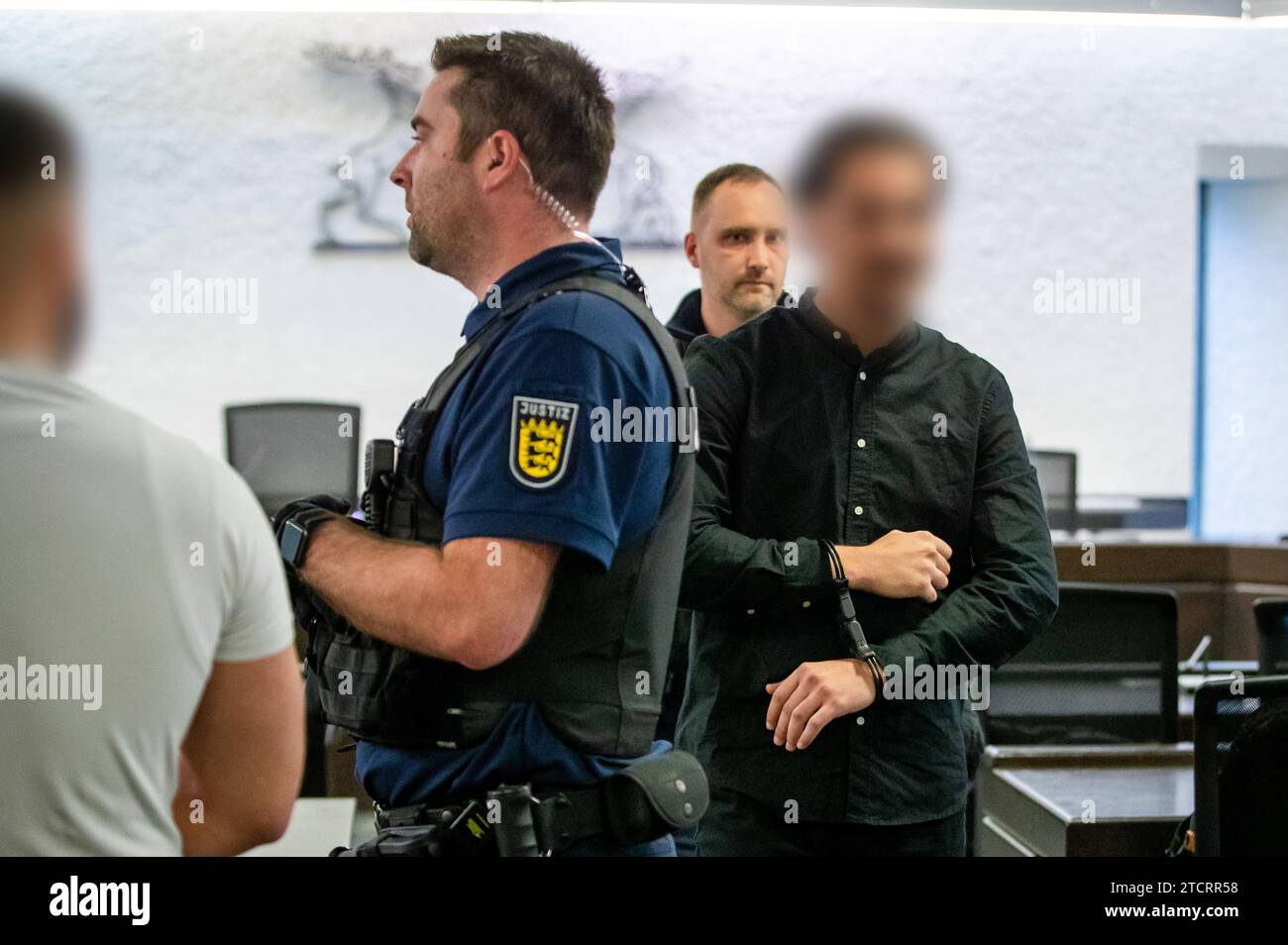 Stuttgart, Germany. 14th Dec, 2023. One of five defendants is led into a courtroom of the district court by a court official. Five men are being tried at Stuttgart Regional Court for attempted manslaughter and causing grievous bodily harm. They allegedly punched and kicked a suspected grenade thrower, who had fled after throwing a hand grenade at a funeral service at a cemetery in Altbach, while he was still in a cab waiting for him and dragged him out of the cab. Credit: --/dpa - ATTENTION: Person(s) has/have been pixelated for legal reasons/dpa/Alamy Live News Stock Photo
