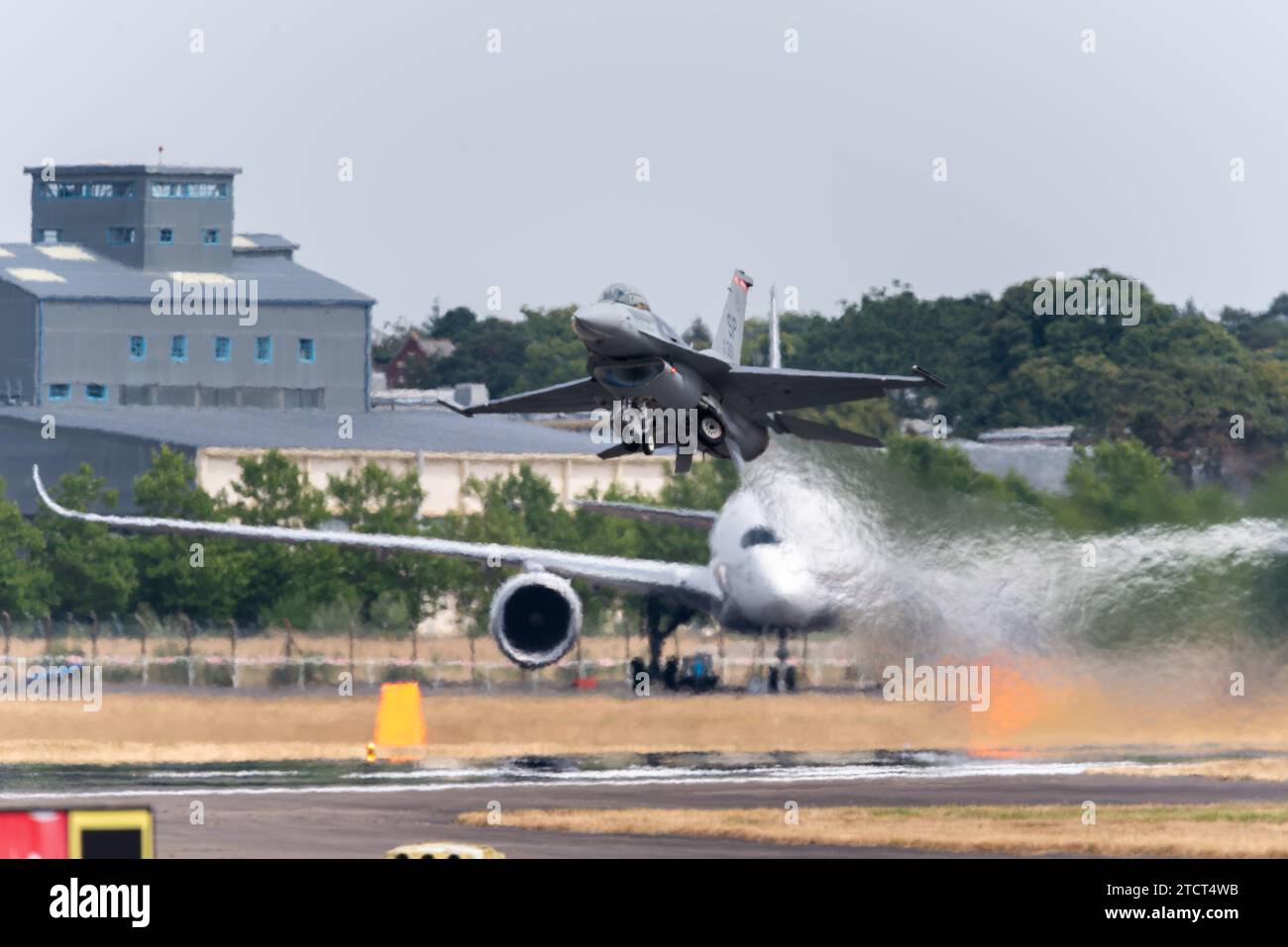 USAF Lockheed Martin F-16C Fighting Falcon taking off from Farnborough Airport with modern airliner and historic buildings in jet efflux heat haze Stock Photo