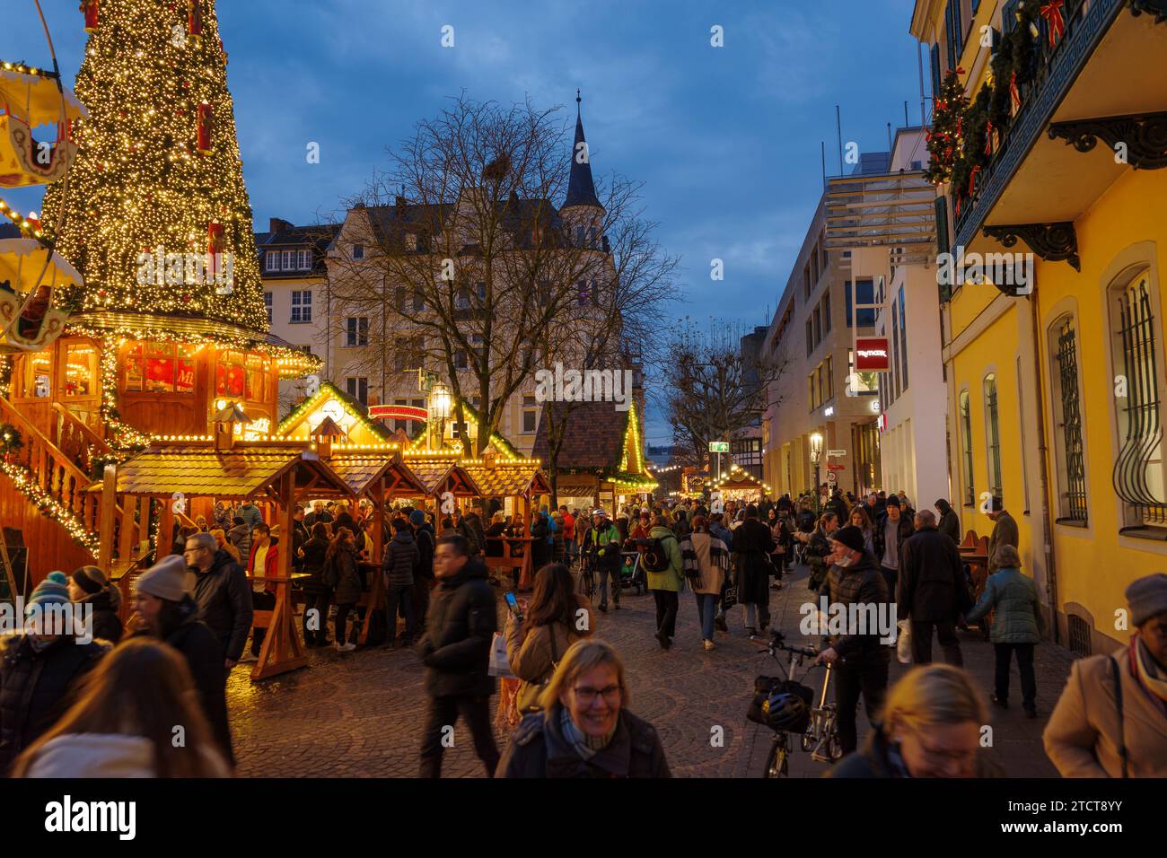 Bonn, Germany - Dec 6, 2023: View of a crowded Christmas market with festive lights, decorated tree, and holiday stalls, with a large illuminated emer Stock Photo