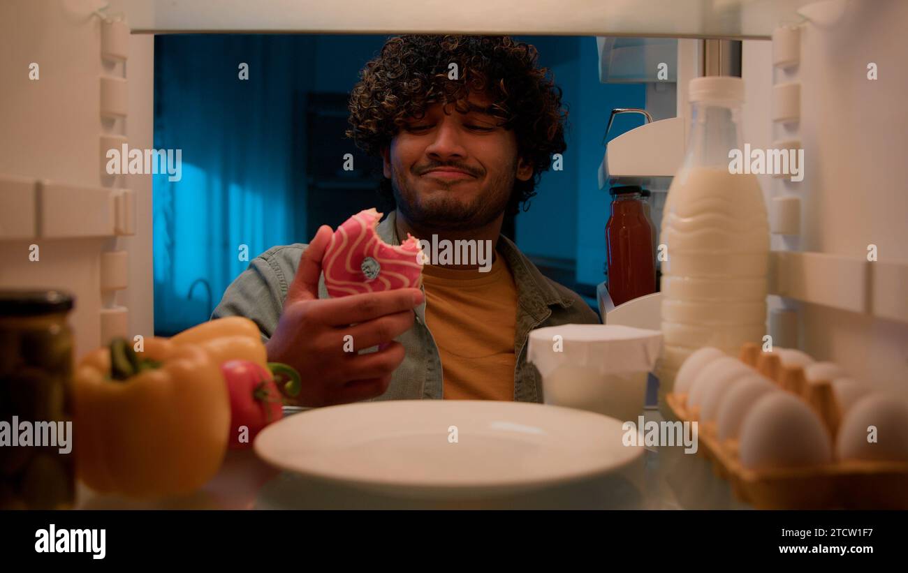 Point of view POV from inside refrigerator hungry Indian man Arabian guy male at night kitchen open fridge want eat take bitten sweet donut smiling Stock Photo