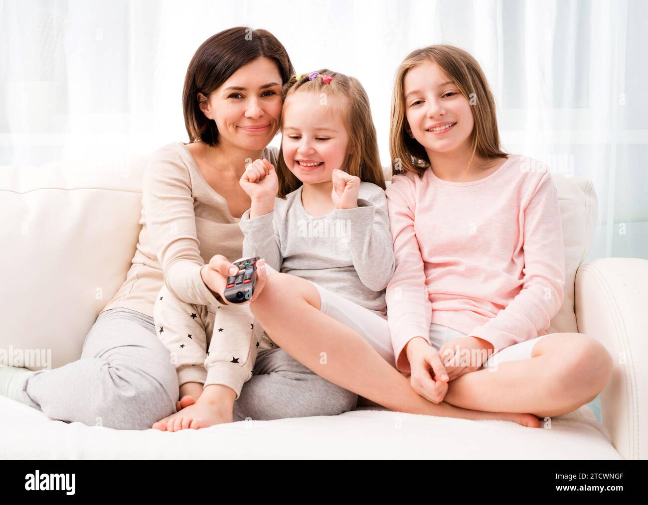 Portrait of mom hugging her cute daughters while sitting on the sofa and watching TV at home. Happy family relaxing on the coach Stock Photo