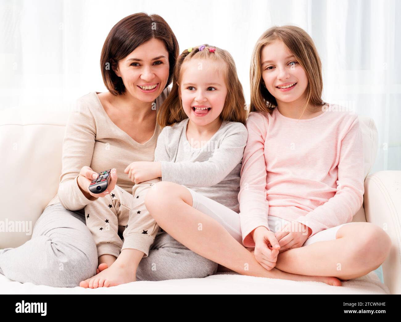 Portrait of mom hugging her cute daughters while sitting on the sofa and watching TV at home. Happy family relaxing on the coach Stock Photo