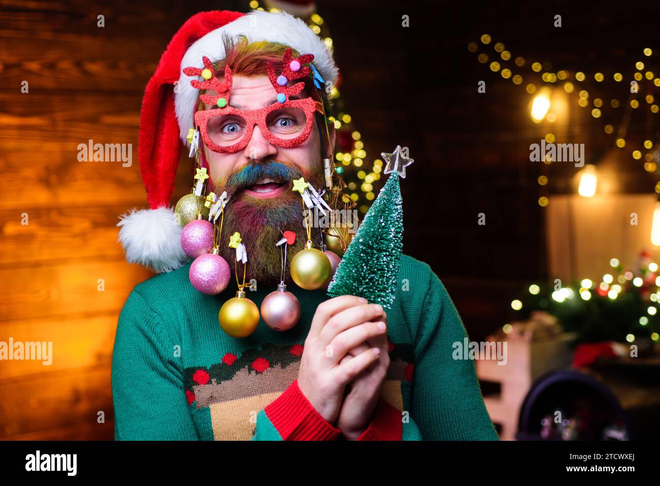 Smiling man with decoration balls in beard with small Christmas tree. Preparation for Christmas or New Year party. Bearded guy in Santa hat and party Stock Photo