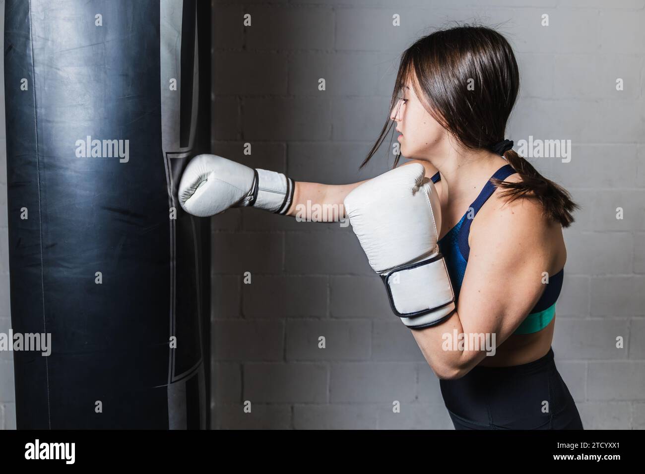Horizontal photo pretty Caucasian teen girl Caucasian, dressed in sportswear and white boxing gloves, on white brick background. Hitting punching bag Stock Photo