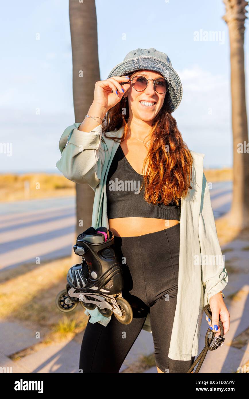 Happy young beautiful Caucasian brunette woman wearing hat and sunglasses standing with inline roller skates against sky in park Stock Photo