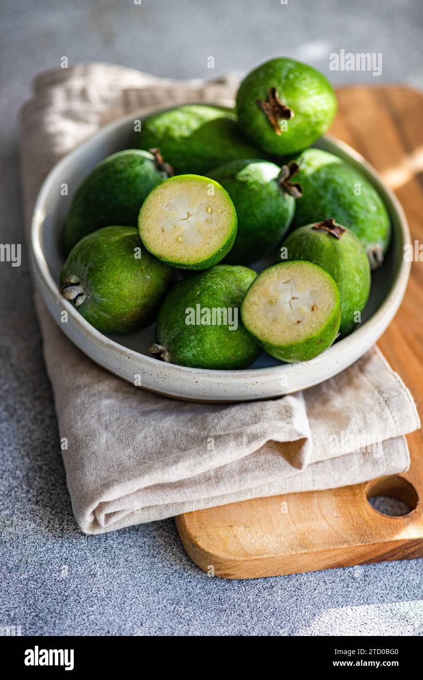 Raw feijoa fruits, also known as pineapple guavas, displayed in a ceramic bowl on a wooden cutting board with a linen cloth. Stock Photo