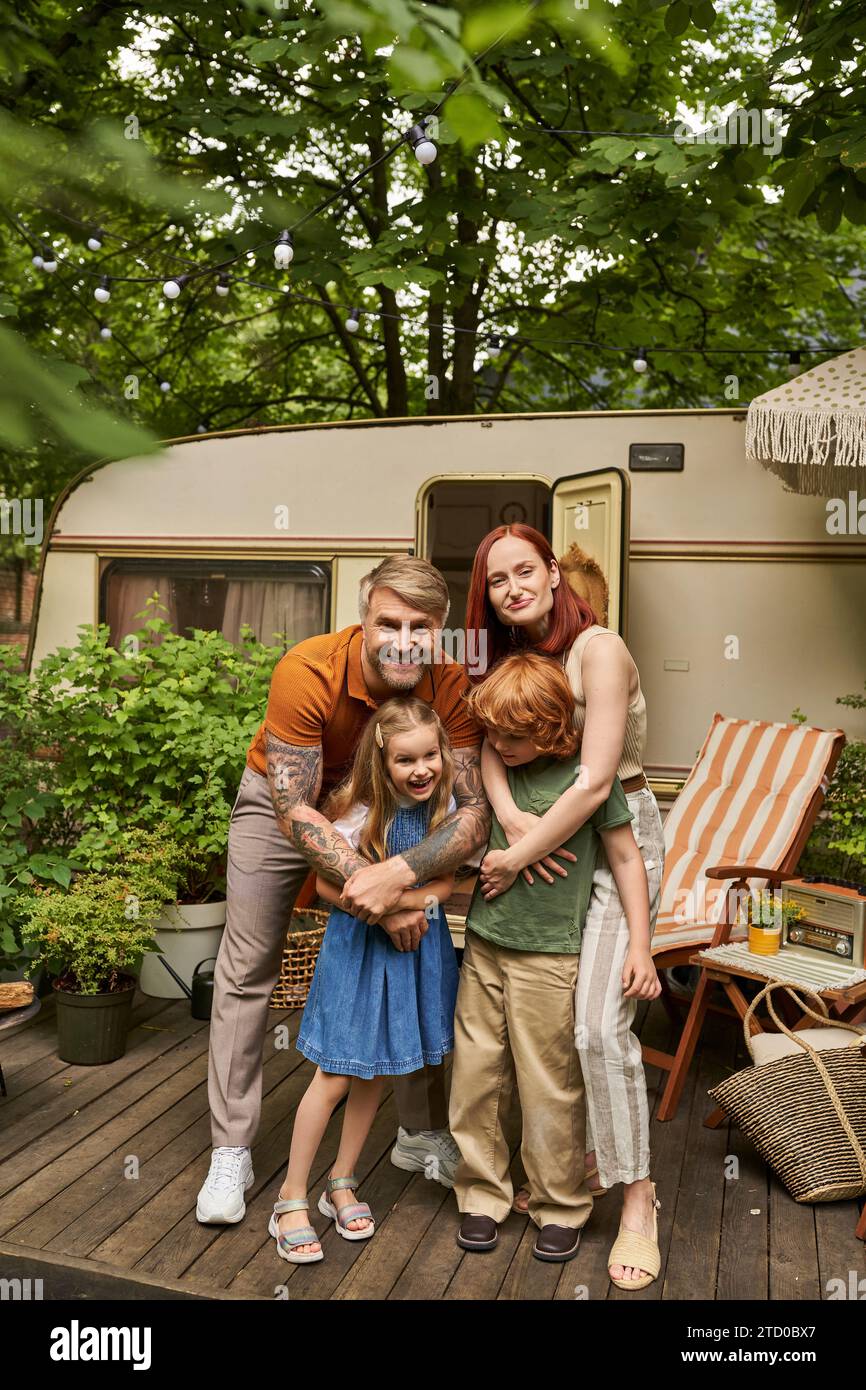 happy parents embracing cheerful children near modern home on wheels in trailer park, family leisure Stock Photo