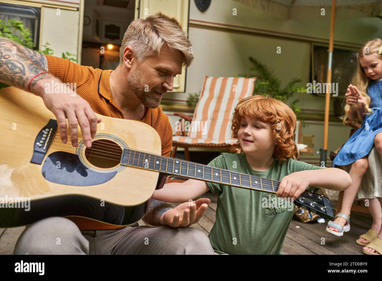 tattooed man teaching redhead son to play acoustic guitar near home on wheels in trailer park Stock Photo