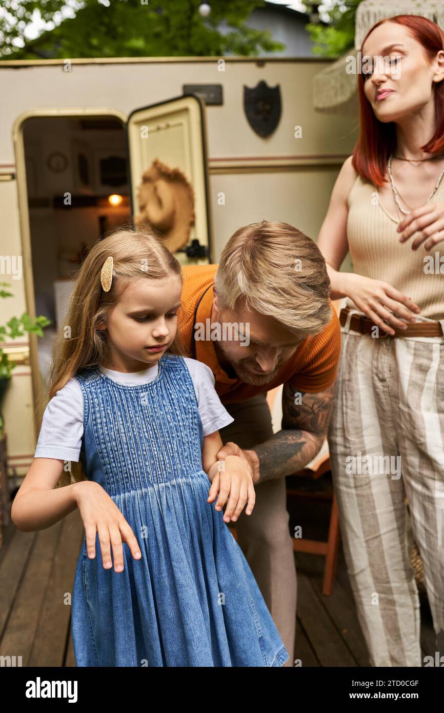 father supporting adorable daughter near wife and home on wheels in trailer park, love and care Stock Photo