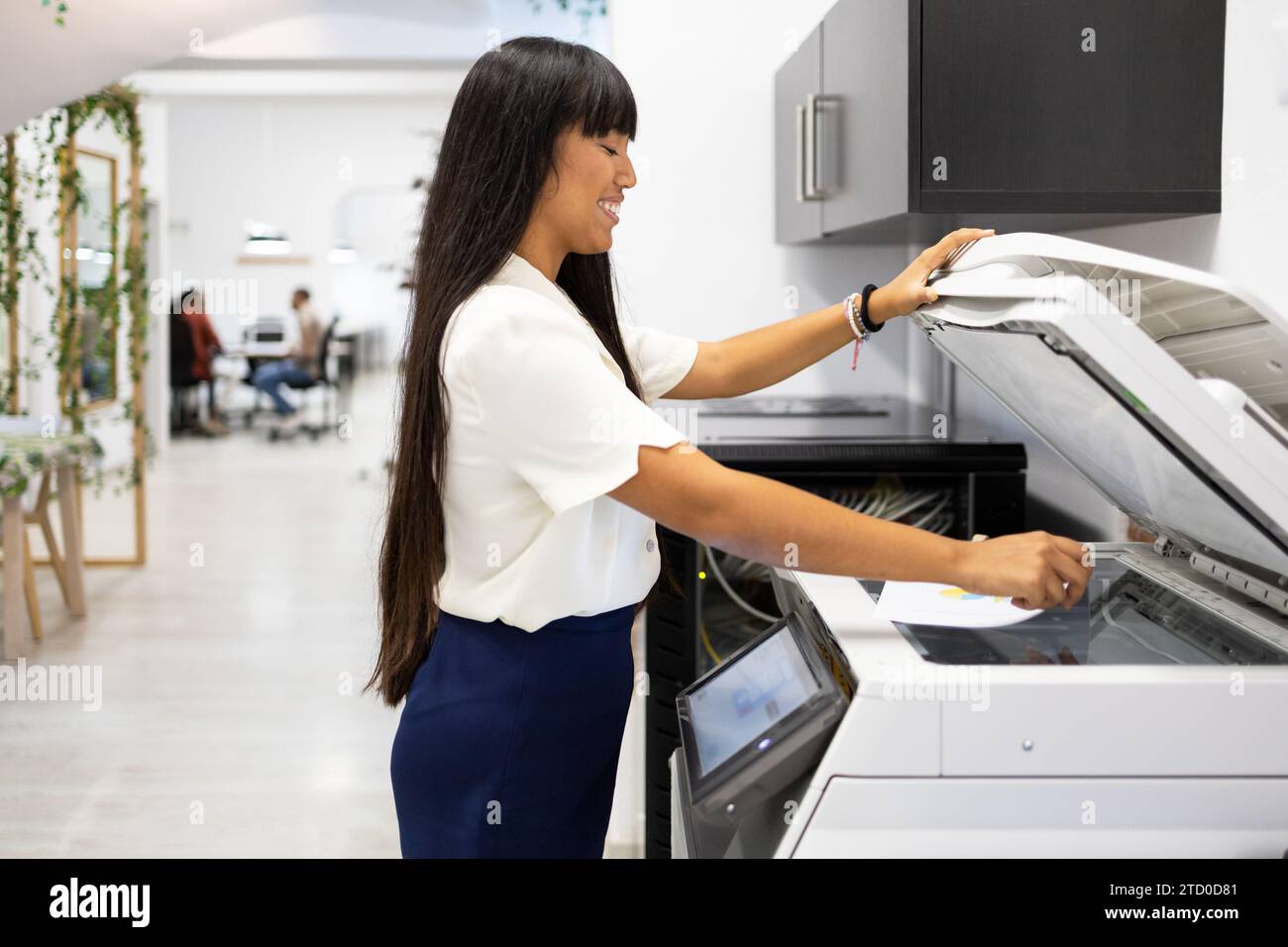 Side view of smiling Asian young businesswoman in casuals with long hair using modern copier while working in creative office Stock Photo