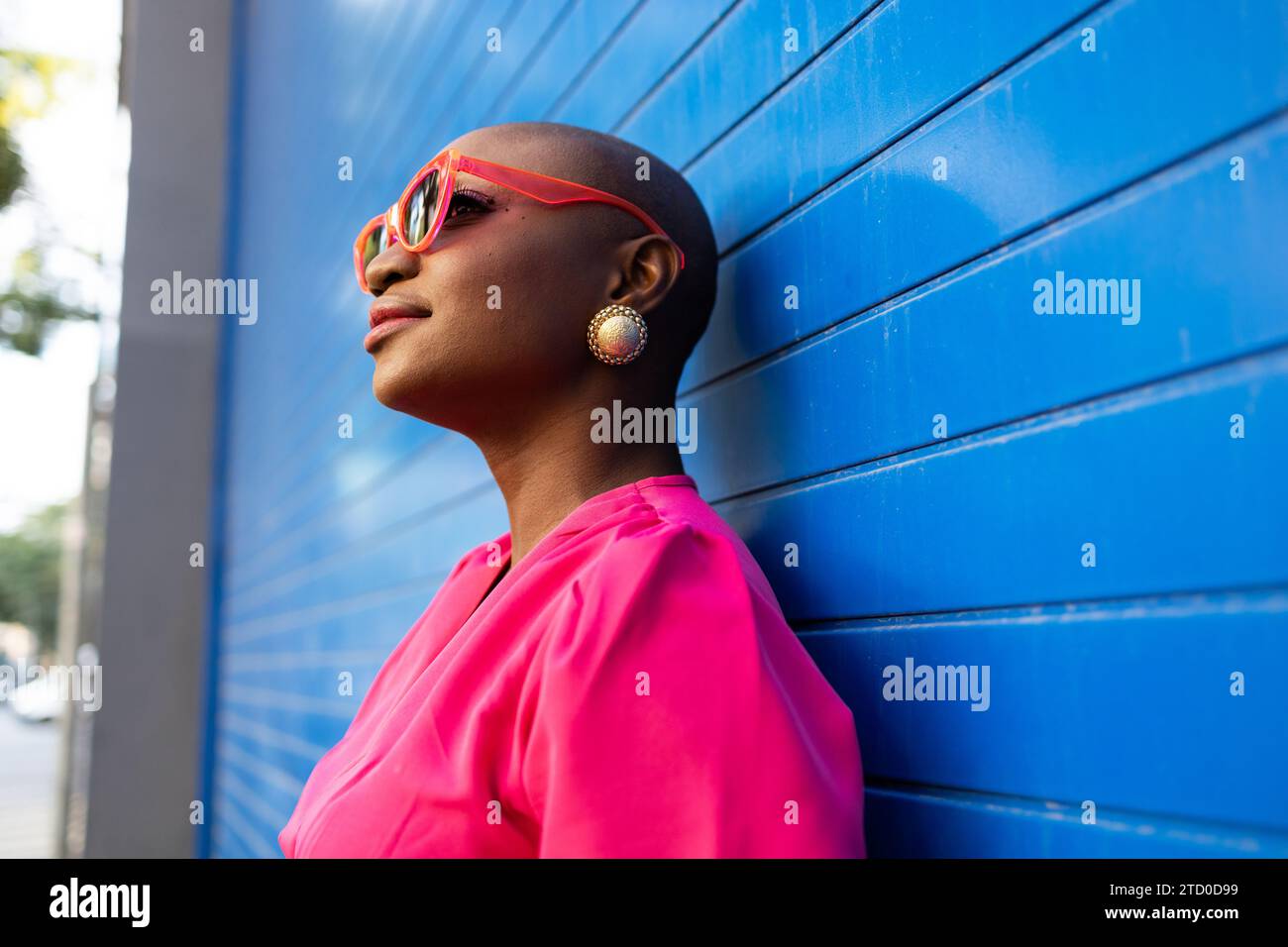 Side view of smiling African American female with shaved head in trendy pink outfit and sunglasses looking away while standing against blue wall Stock Photo