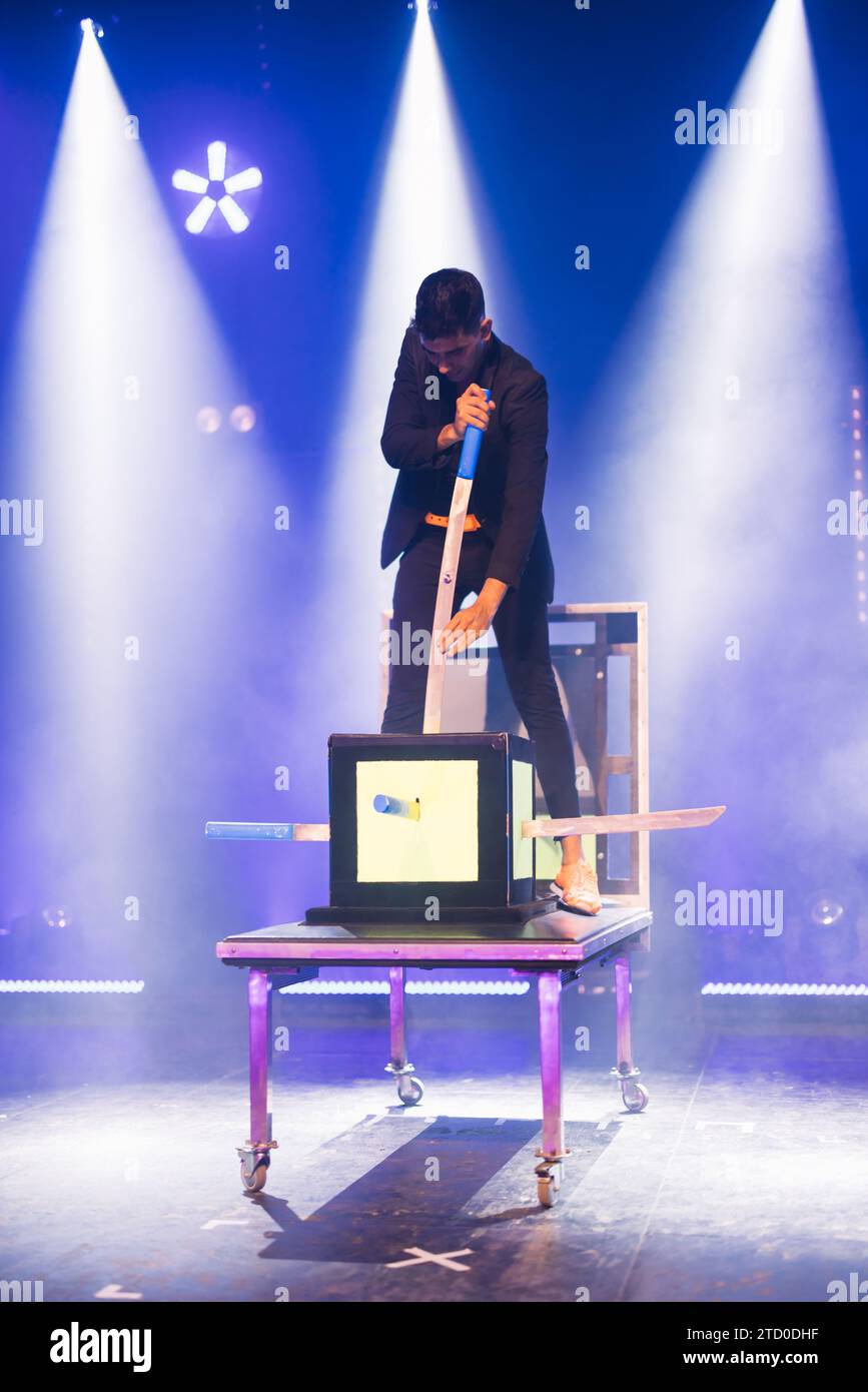 Professional male magician on top of table showing magic trick with a sword and glass boxes on stage with smoke and glowing spotlights during performa Stock Photo