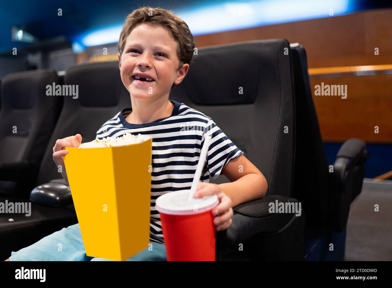 Cheerful cute boy holding tasty popcorn bucket and disposable coke cup while sitting on black seat and watching movie at theater during weekend Stock Photo