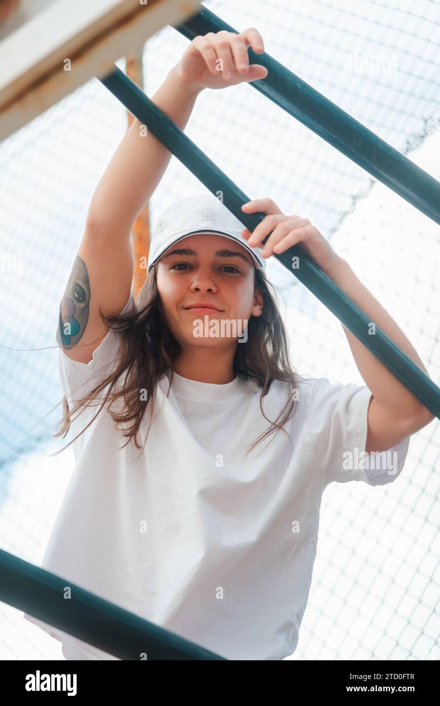 A young woman with a tattoo on her arm poses casually behind a green metal fence, looking relaxed and confident in a simple white tee Stock Photo