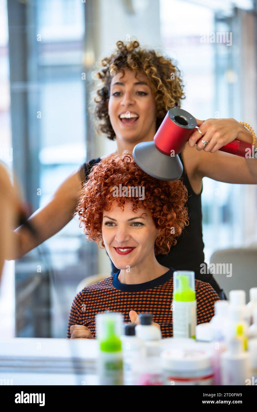 Professional hair stylist using a blow dryer on a smiling woman with vibrant curly red hair in a modern salon setting. Stock Photo
