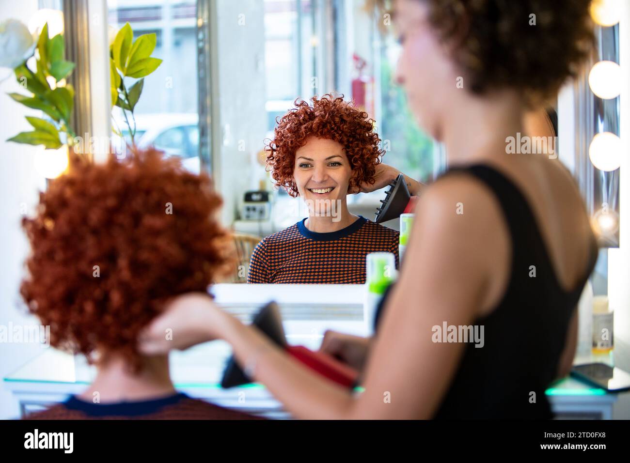A cheerful woman with curly red hair is styling her hair using a straightener in a bright, modern hair salon, with reflections in the mirror. Stock Photo