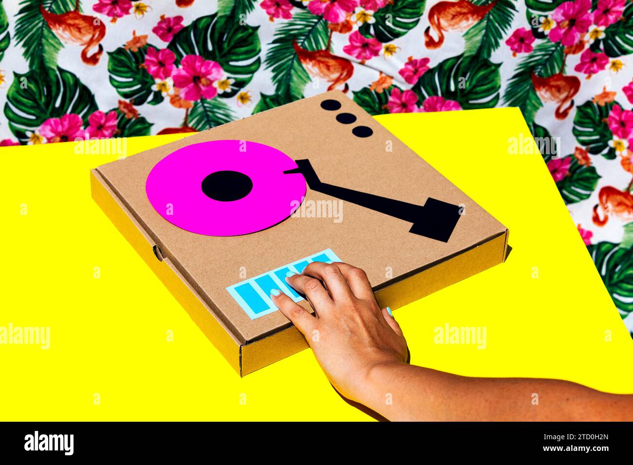 Crop hand of anonymous woman playing antique turntable made with cardboard box on yellow table over floral background Stock Photo