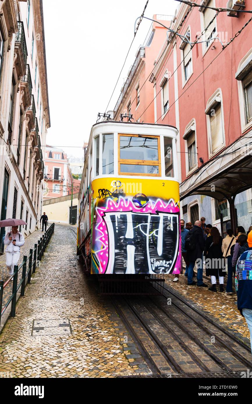 Graffiti covered Tram 28 procedes up a steep incline on Rua da Conceicao past waiting passengers. Lisbon, Portugal Stock Photo