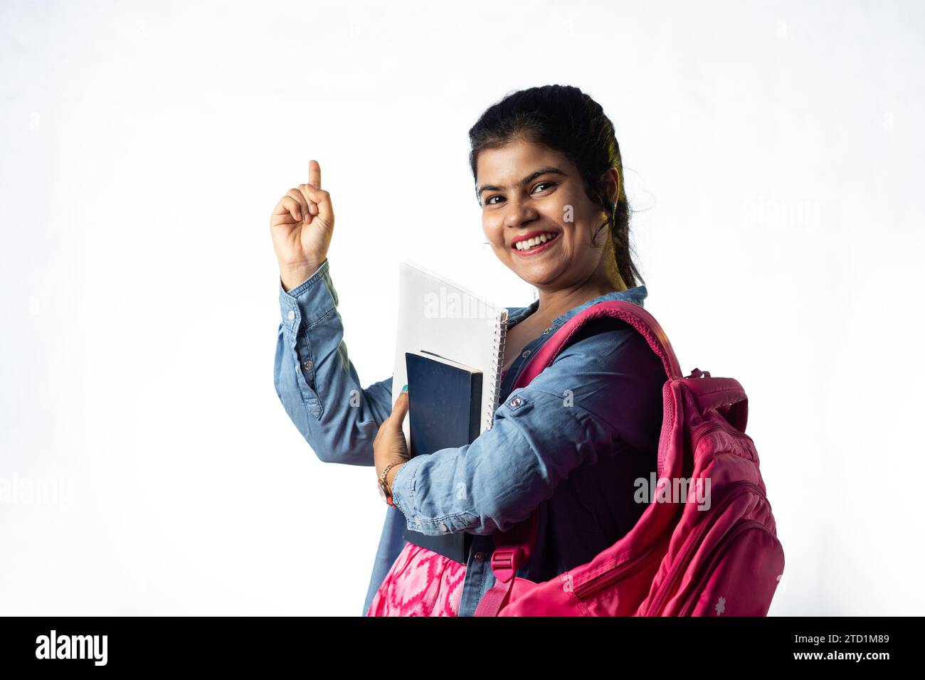A pretty young Indian girl with books in hand pointing with smiling ...
